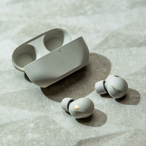 A high-end, clean product shot of off-white wireless earbuds and their charging case resting on a textured light gray surface.