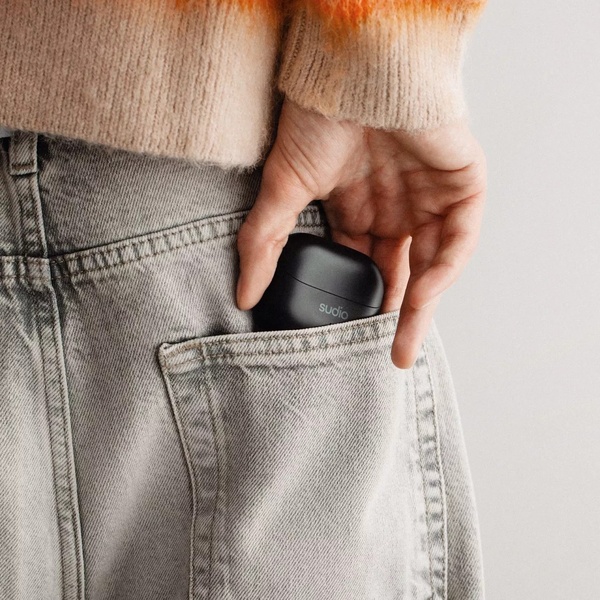 A close-up shot of a hand placing a sleek, black wireless earbuds case into the back pocket of grey jeans.
