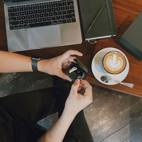 A high-angle shot of a clean desk featuring a person holding wireless earbuds in a charging case next to a laptop and latte.