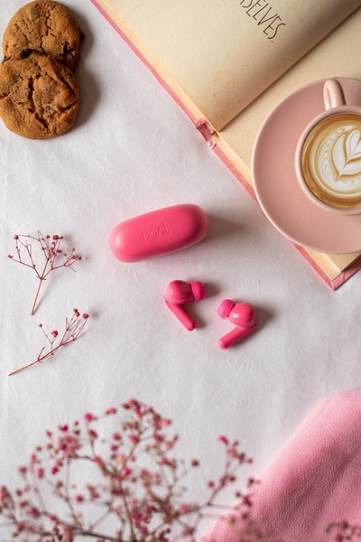 This creative displays a pair of pink wireless earbuds placed on a clean white surface alongside a coffee cup, cookies, and floral elements.