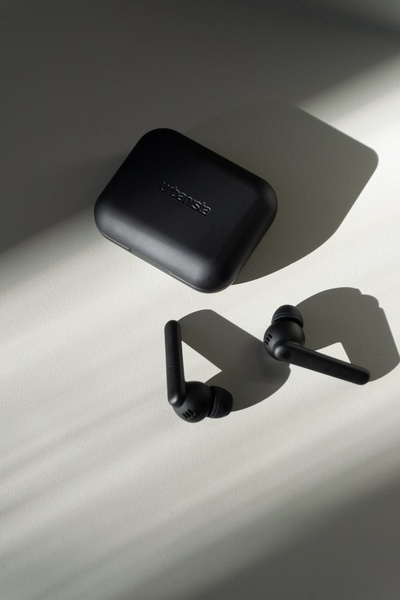 A high-contrast, studio-style shot of black wireless earbuds and their charging case on a light neutral background, utilizing dramatic natural light and shadow.
