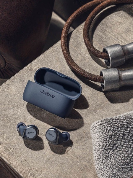 A high-end, clean product shot of Jabra earbuds and their charging case arranged on a textured wooden surface next to workout gear.