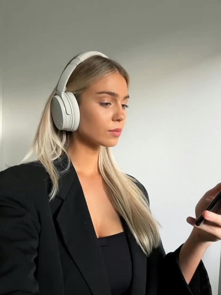A close-up portrait of a woman wearing modern white wireless headphones while looking at her smartphone in a professional setting.