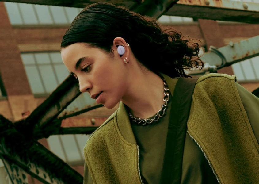 A portrait-focused, moody lifestyle shot of a woman wearing wireless earbuds in an urban industrial setting.