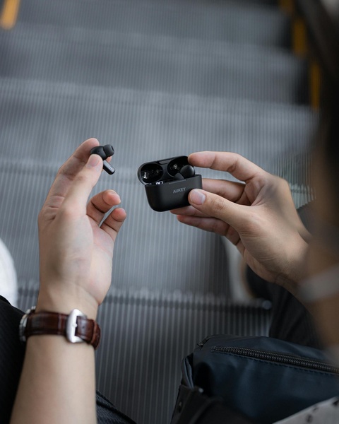A close-up, first-person perspective shot of a user holding wireless earbuds next to their charging case while on an escalator.