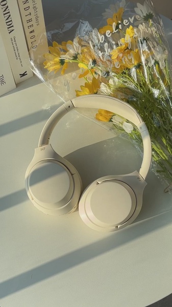 A clean, high-angle product shot of beige wireless headphones placed next to a bouquet of yellow flowers, utilizing soft, natural morning sunlight.