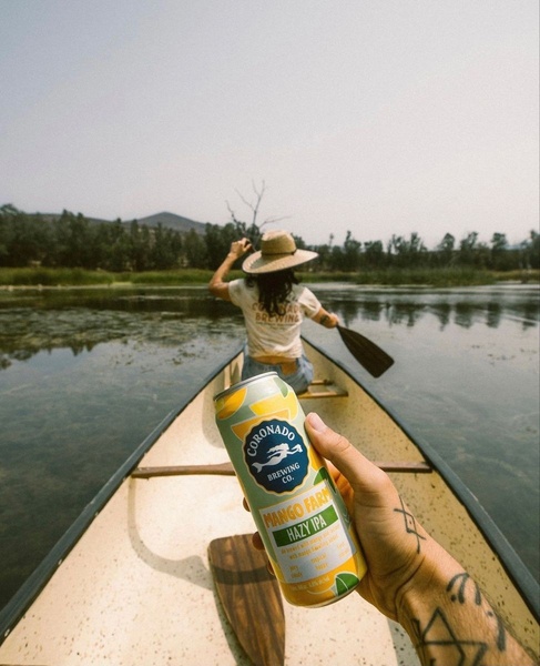 A first-person view of someone holding a Hazy IPA can while canoeing on a calm, scenic lake.