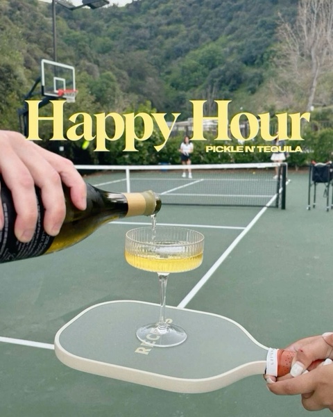 A high-end lifestyle shot featuring a pickleball paddle serving as a drink coaster on a tennis court, capturing a luxury social atmosphere.