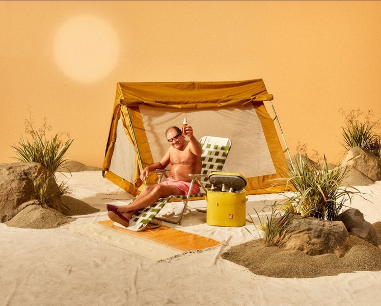 A man relaxes in a camping chair inside a tent located in a desert landscape, holding a cold drink, evoking a feeling of isolation and refreshment.