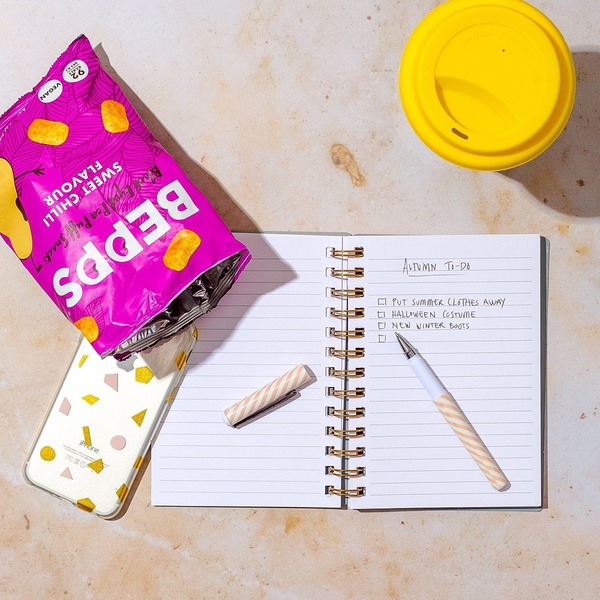 A top-down, minimalist desk scene featuring a bag of sweet chilli snacks next to an open notebook containing an autumn to-do list.