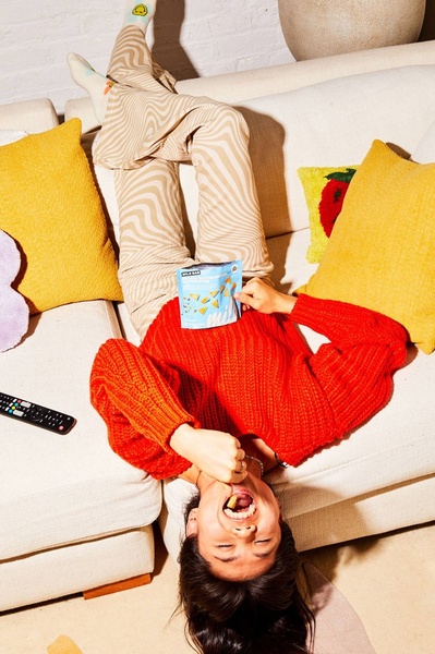 A high-angle, relatable shot of a person relaxing on a sofa, eating snacks directly from the bag while upside down.