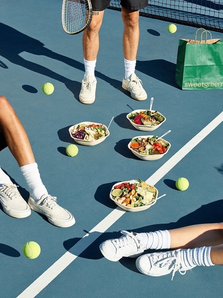 A high-angle, top-down shot of a tennis court surface featuring multiple salad bowls and tennis gear, blending active living with fresh food.