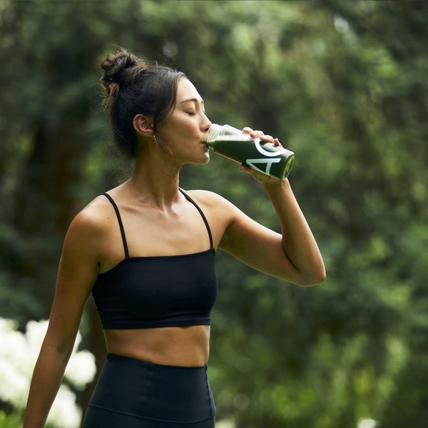A clean, outdoor lifestyle shot of a woman consuming a green health drink in a natural setting.