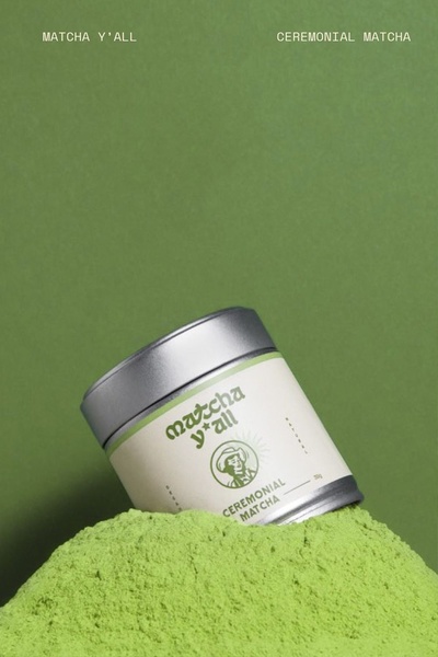 A clean, eye-catching product shot of a ceremonial matcha tin resting in a mound of bright green matcha powder against a solid earth-tone background.