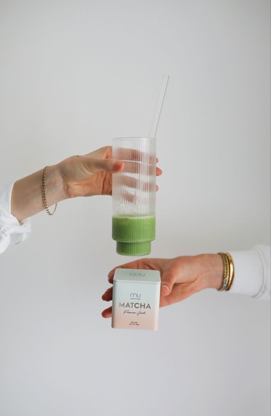 A clean, high-key studio shot featuring a woman's hands presenting a glass of iced matcha alongside its premium product packaging.