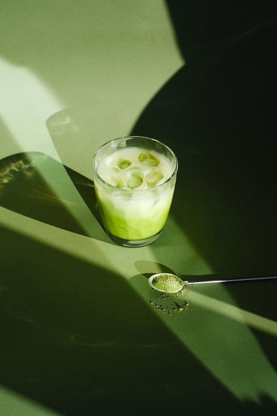 A high-quality lifestyle product shot of a matcha latte in a glass, with a spoon of powder in the foreground, set against a deep green backdrop with dramatic light play.