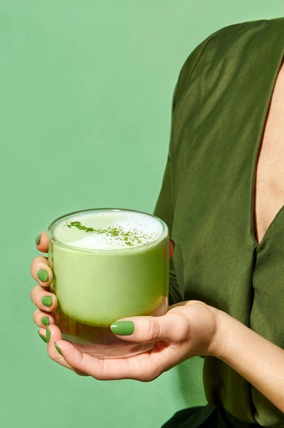 A close-up, high-quality portrait shot of a person holding a glass of frothy green matcha tea against a solid light green background.