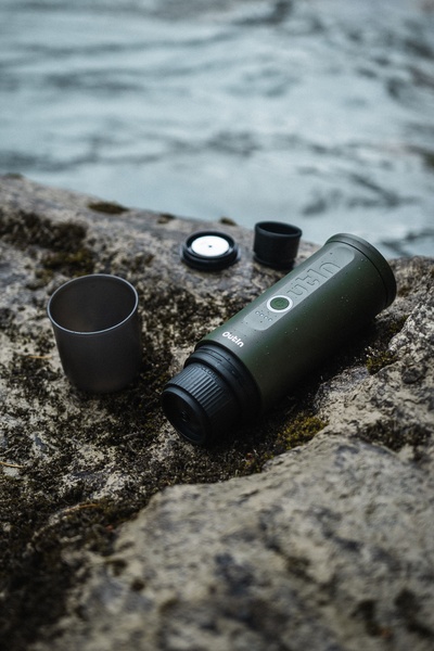 A close-up shot of a portable espresso maker sitting on a natural rock surface by the water, emphasizing portability and lifestyle.