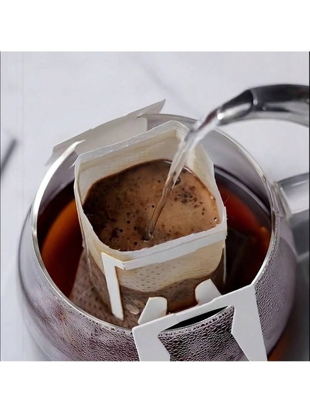 A close-up shot focusing on the tactile experience of pouring hot water over a single-serve drip coffee bag inside a glass vessel.