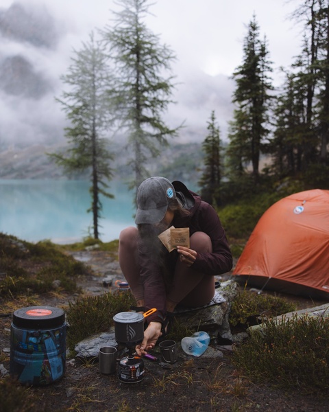 A candid shot of a hiker preparing instant coffee with a portable stove in a misty, mountain lakeside setting.
