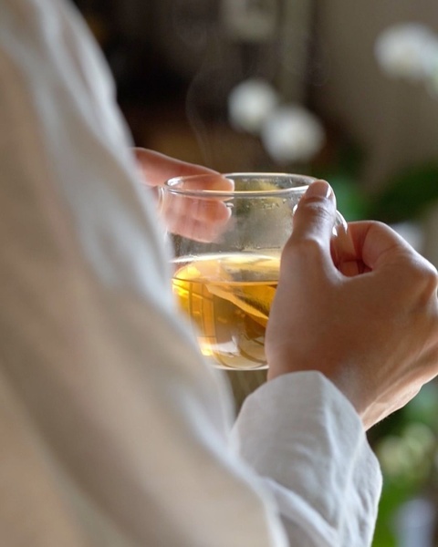 A close-up, warm-toned shot of hands holding a clear glass of steaming hot tea in a quiet, serene environment.