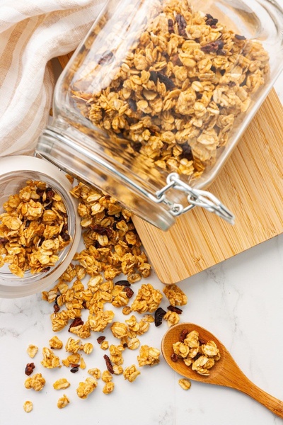 A high-quality top-down shot of organic granola pouring from a clear glass jar onto a clean white surface with a wooden spoon.