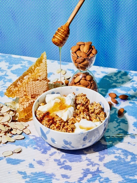 A high-end studio shot of a granola bowl featuring honey being drizzled, surrounded by honeycomb and almonds on a blue patterned background.