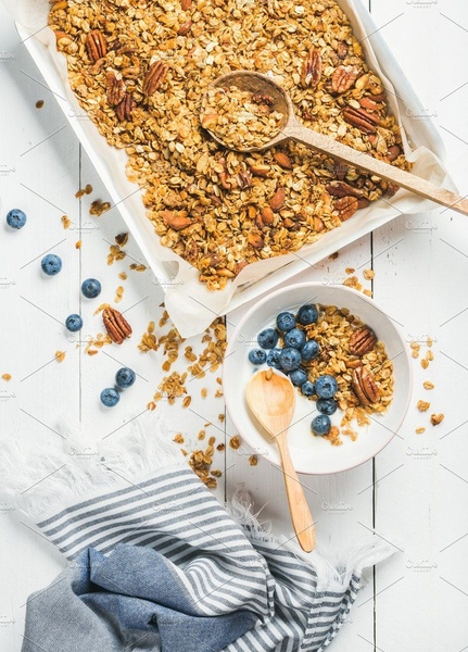 A top-down view of homemade granola on parchment paper next to a bowl with granola and blueberries, set against a bright wooden surface.