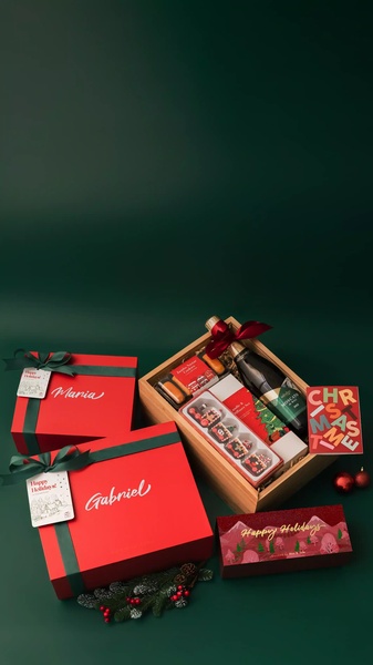 A high-end, clean product photography layout featuring curated holiday gift boxes with premium wine and seasonal treats, set against a deep green backdrop.