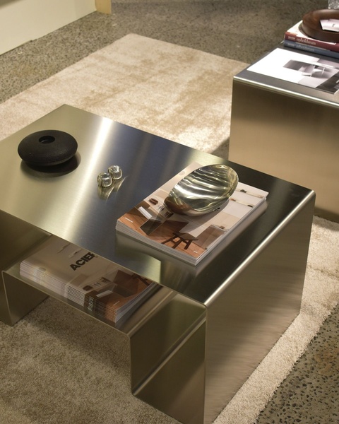 A high-end, clean product hero shot featuring a metal coffee table staged with curated lifestyle objects like books and metallic bowls.