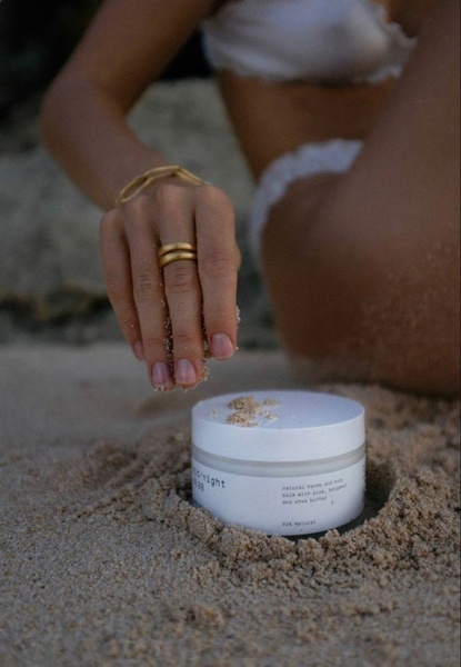 A close-up aesthetic shot of a hand and body balm jar placed in the sand at a beach, held by someone in beachwear.