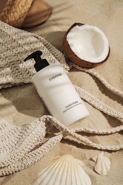 A high-angle studio photograph of a body and hand cream bottle resting on white sand, accompanied by a halved coconut, seashells, and woven accessories.