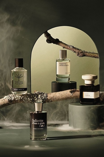 A high-end product hero shot featuring four distinct perfume bottles arranged amidst organic tree branches and soft, atmospheric smoke.