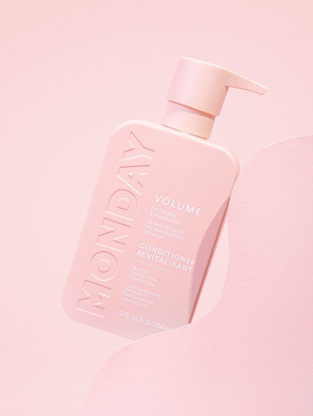 A clean, studio-lit photograph of a pastel pink hair conditioner bottle positioned on a textured surface of the same color.
