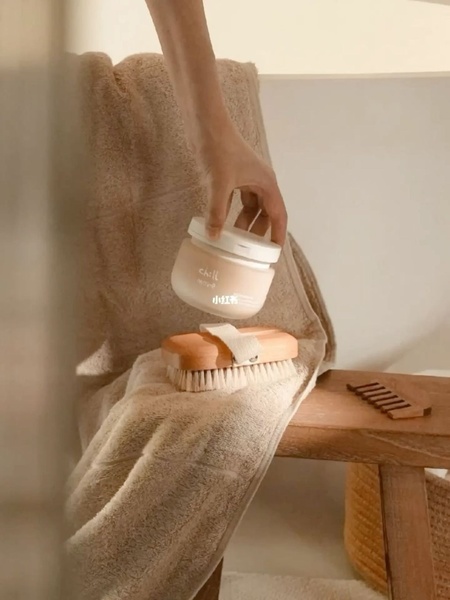 A soft-lit, high-quality product shot featuring a bath brush and a jar of cream on a wooden stool, conveying a peaceful wellness experience.