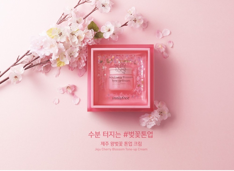 A high-end product hero shot featuring the cream centered in a pink box with cherry blossom branches, emphasizing a natural, botanical, and fresh aesthetic.
