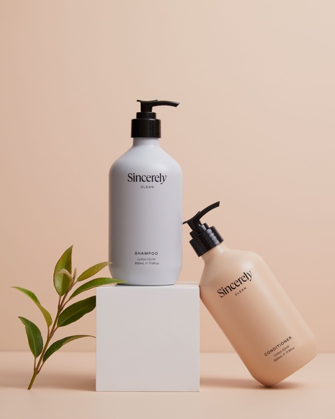A clean, studio-lit photograph featuring two shampoo and conditioner bottles on a geometric pedestal with a natural botanical accent.