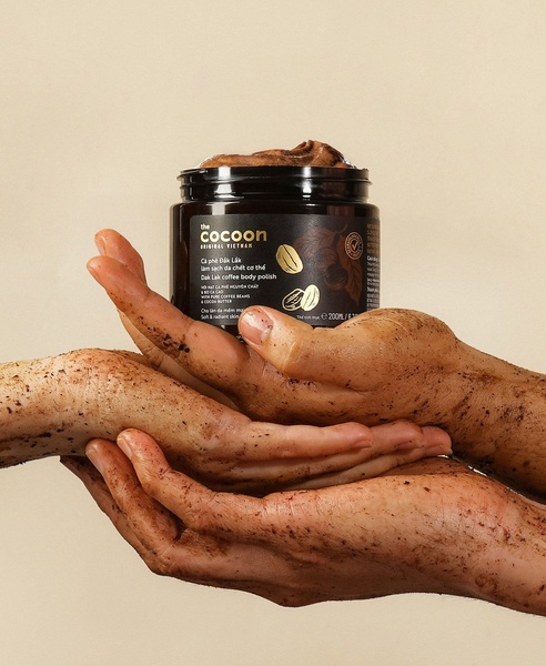 A close-up, high-quality product shot of a Coffee Body Polish jar being held by hands covered in the product, emphasizing texture and natural ingredients.