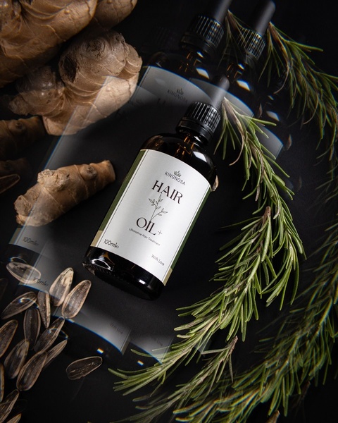 A high-end product photography shot of Kingnosa Hair Oil+ surrounded by raw ingredients like ginger, rosemary, and seeds on a dark, reflective surface.