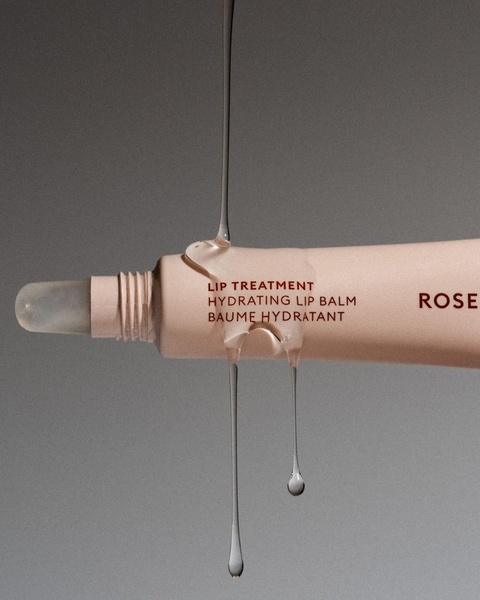 A high-end, minimalist product hero shot featuring a clear lip treatment gel dripping from a tube against a neutral, clean backdrop.