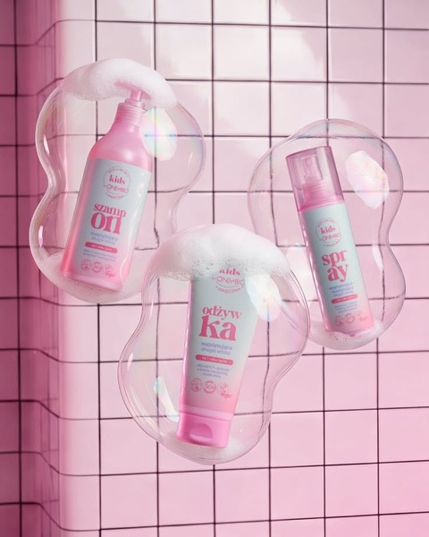 A high-key studio shot of shampoo, conditioner, and spray products encased in soap bubbles against a light pink grid background.