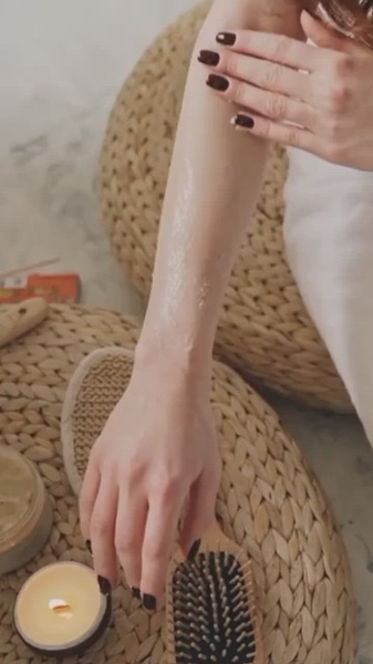 A close-up aesthetic shot of a person applying body scrub to their arm, accompanied by a burning candle and a hairbrush on a wicker surface.