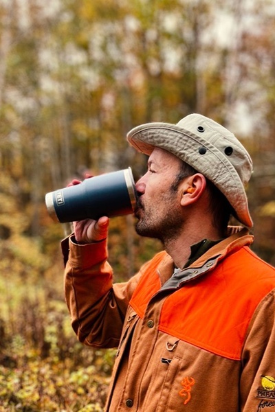 A high-quality lifestyle shot of a man in hunting gear drinking from a branded insulated tumbler in a natural, wooded environment.