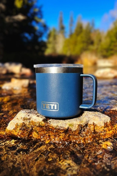 A high-quality image of an insulated blue mug placed on a rock in a flowing stream, emphasizing durability and outdoor utility through natural lighting.