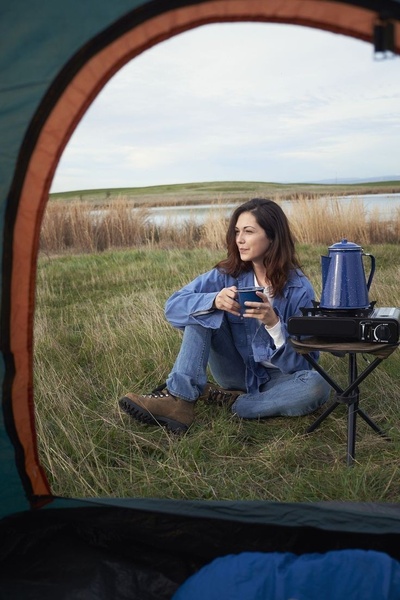 A first-person perspective shot from inside a tent showing a person enjoying a hot drink next to a portable camping stove in a serene, natural environment.