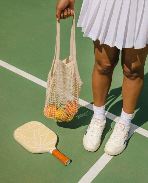 This creative showcases a stylized pickleball paddle alongside a mesh bag of produce on a court, emphasizing a leisure-focused brand aesthetic.