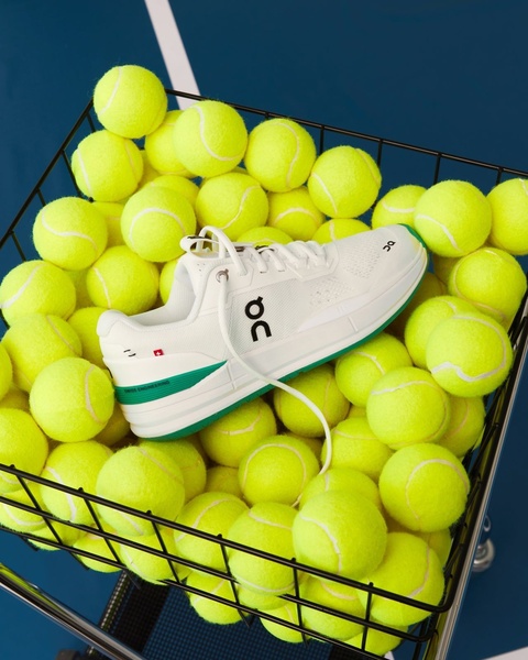 A high-angle, clean product shot of a white running shoe centered within a ball hopper filled with neon tennis balls.