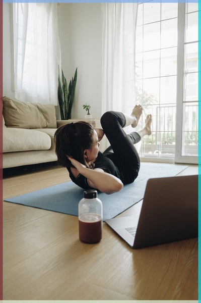 A high-angle shot of a woman performing core exercises on a blue yoga mat in a brightly lit living room, with a protein drink and laptop nearby.