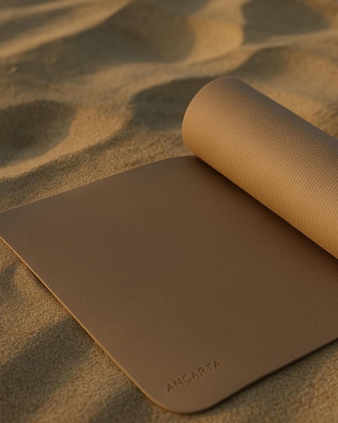 A high-end, clean shot of a yoga mat placed on sand, utilizing natural lighting and soft shadows to communicate product texture and quality.