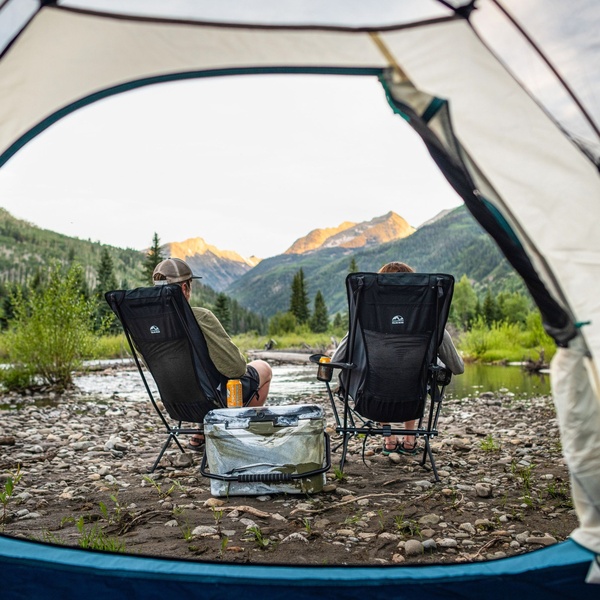 A high-quality 1:1 image showing a first-person perspective from inside a tent looking out at two people sitting in folding chairs in a scenic mountain valley.